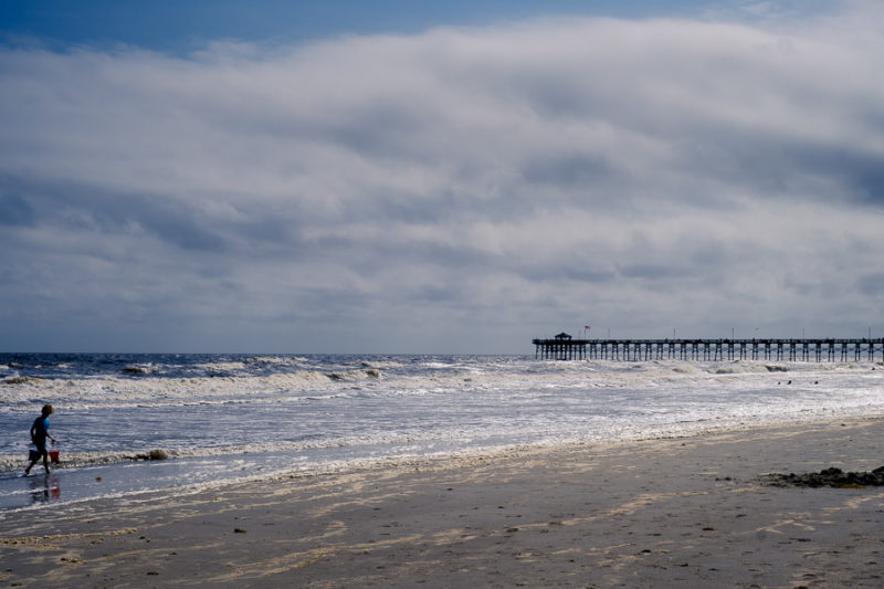 Beach scene with a pier in the distance and cloudy sky. There is also a small child in the left lower corner of water carrying buckets from the ocean to build a sand castle.  